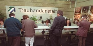 Menschen stehen mit Formularen an einem Bürothresen, hinter dem Sachbearbeiterinnen sitzen. Im Hintergrund an der Wand steht das Wort "Treuhandanstalt".
