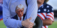 Ein Mann trägt ein helbblaues Shirt mit dem Porträt von Donald Trump, hinter ihm steht ein Mann in einem Shirt in den Farben der US-Flagge
