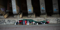 Ein Mensch liegt in einem grünen Schlafsack auf grauem Steinboden neben Brückenpfeilern.