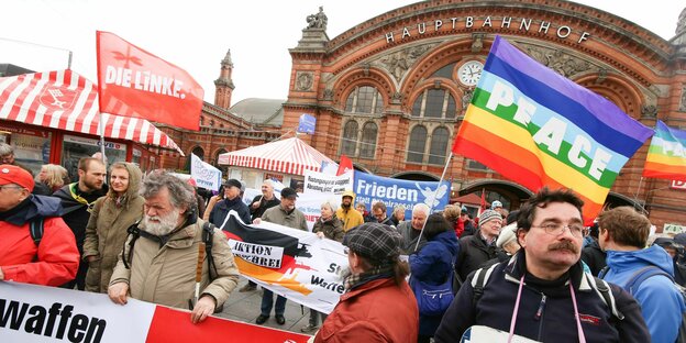 Teilnehmende beim Ostermarsch 2017 mit Plakaten und Fahnen vor dem Bremer Hauptbahnhof. Zu sehen sind unter anderem eine "Peace"-Flagge in Regenbogenfarben, ein Transparent mit der Friedenstaube, sowie ein Spruchband der DKP und eine Fahne der Partei Die Linke.