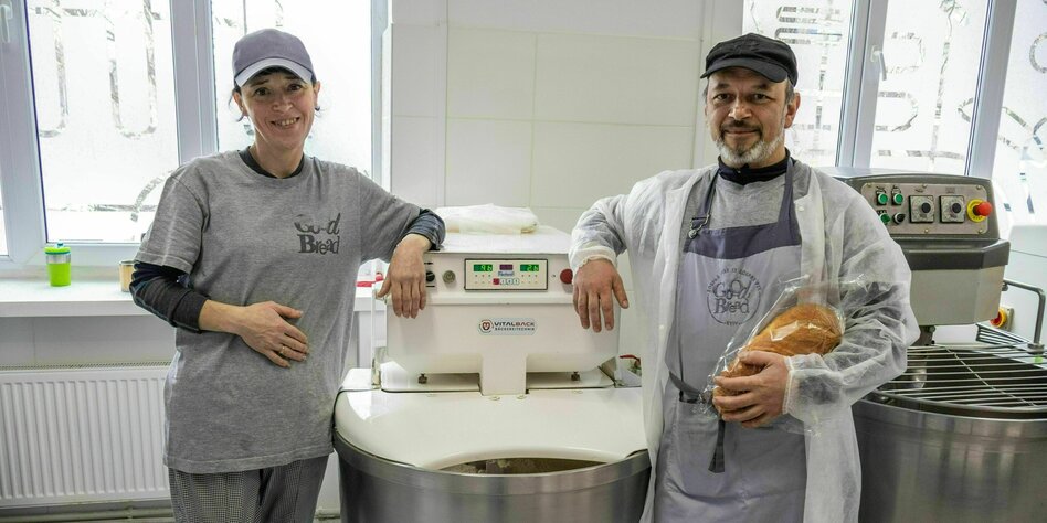 Ein Mann und eine Frau stehen lächelnd vor einer Maschine zum Brotbacken, der Mann hat ein Brot in der Hand