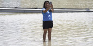 Eine Frau steht auf einer knöchelhoch überfluteten Straße und macht ein Selfie