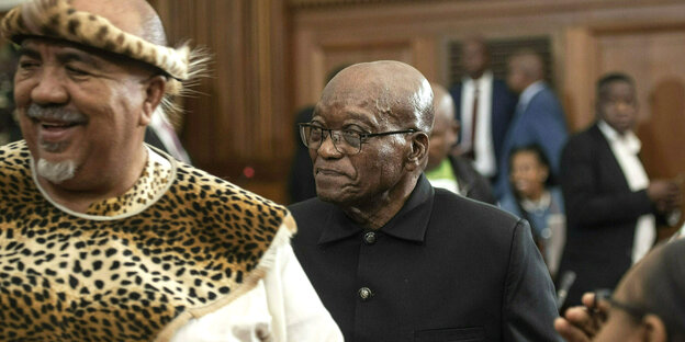 Jacob Zuma geht lächelnd durch einen Raum, vor ihm steht ein Mann, mit Leoparden-Kopfbedekcung und einer Leoparden-Schärpe