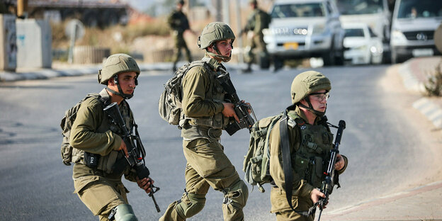 Drei Soldaten in voller Montur und mit Waffen überqueren eine Straße - im Hintergrund hindern weitere Soldaten Fahrzeuge an der Weiterfahrt