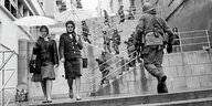 Zwei Frauen mit Regenschirmen gehen eine Treppe hinunter, hinauf gehen Soldaten mit Gewehren